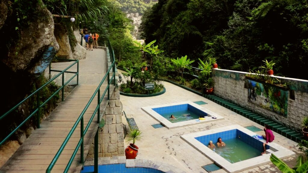 Pozas de los baños termales de Aguas Calientes rodeadas de vegetación de selva, con bañistas y una pasarela de madera