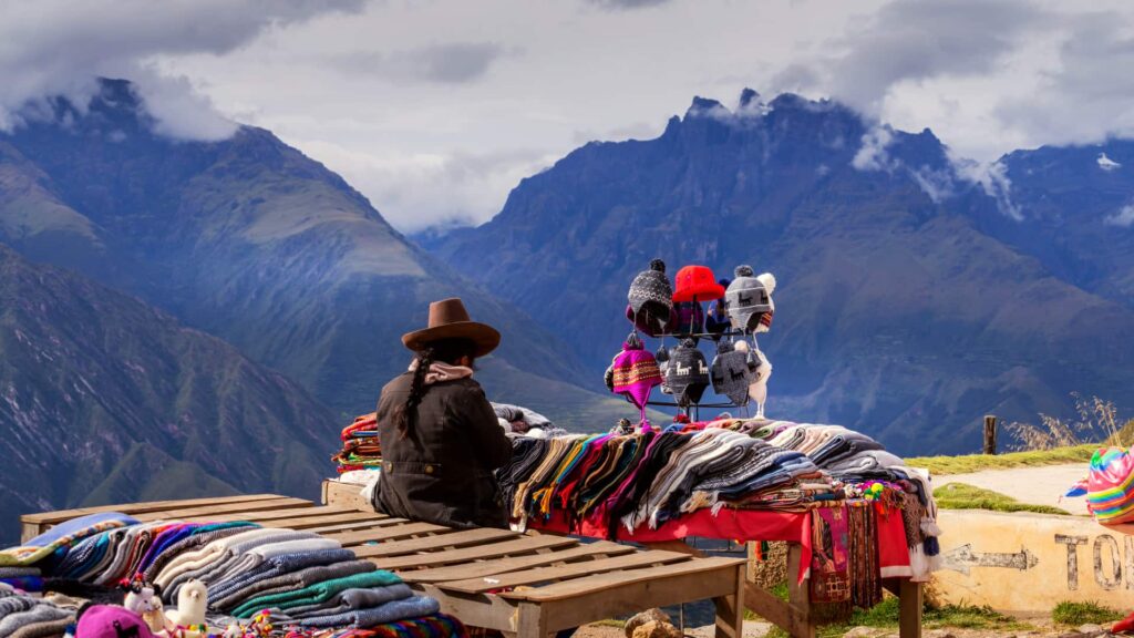 Tejedora con sombrero acomoda textiles andinos en un mirador frente a las montañas de Cusco