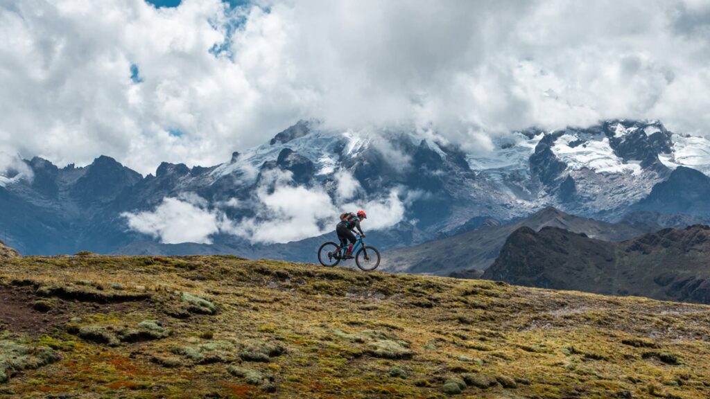 Ciclista de montaña recorriendo un sendero en lo alto de los Andes, con el imponente glaciar del Apu Veronica cubierto de nubes al fondo.