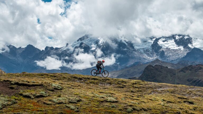 Ciclista de montaña recorriendo un sendero en lo alto de los Andes, con el imponente glaciar del Apu Veronica cubierto de nubes al fondo.