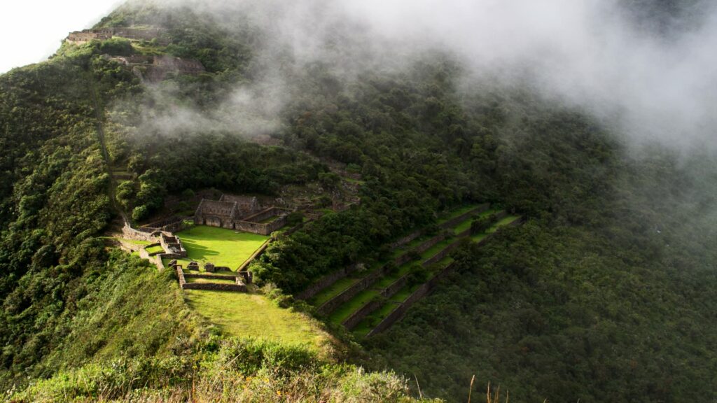 La plaza principal y las ruinas de Choquequirao emergiendo de una densa niebla matutina sobre la verde montaña de los Andes peruanos.