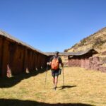 Excursionista con mochila caminando entre los imponentes muros de piedra y adobe del sitio arqueológico inca de Huchuy Qosqo, Perú.