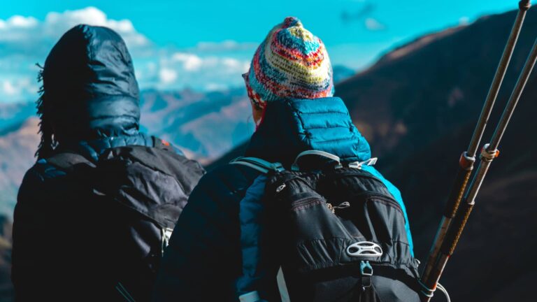 Dos excursionistas de espaldas, equipados con mochilas y gorros, hacen una pausa para admirar el vasto paisaje de montañas en lares trek en Perú.