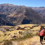 Dos excursionistas caminando por un sendero andino con vistas a las antiguas ruinas incas de Huchuy Qosqo en el Valle Sagrado.