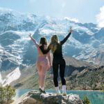 Dos mujeres celebran con los brazos en alto al llegar a la Laguna Humantay, de aguas turquesas, con el imponente nevado Salkantay de fondo.