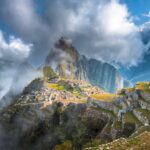 La icónica ciudadela inca de Machu Picchu con la montaña Huayna Picchu de fondo, envuelta en nubes y niebla mística al amanecer.