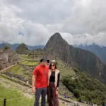Pareja de viajeros en la vista clásica de Machu Picchu con Huayna Picchu entre nubes